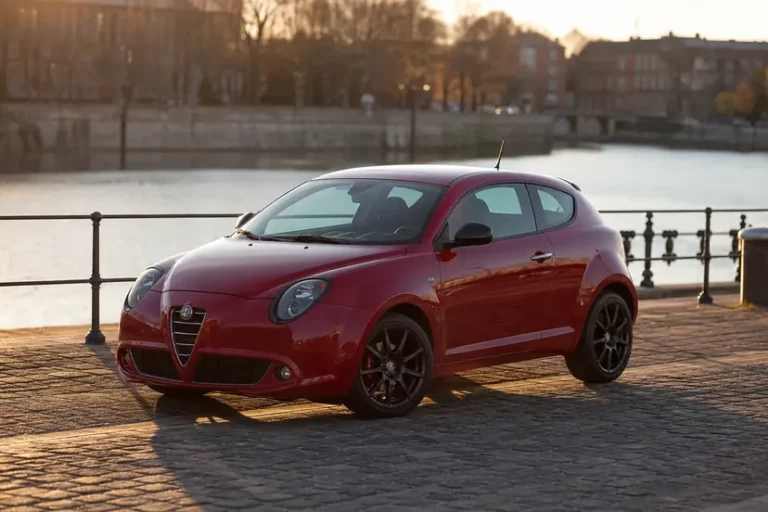 Alfa Romeo MiTo rouge foncé vue de trois quarts avant, arrêtée moteur coupé le long d'un quai pavé au bord d'un canal, en fin de journée