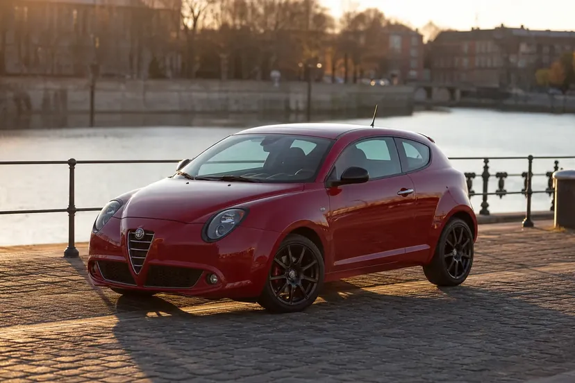 Alfa Romeo MiTo rouge foncé vue de trois quarts avant, arrêtée moteur coupé le long d'un quai pavé au bord d'un canal, en fin de journée