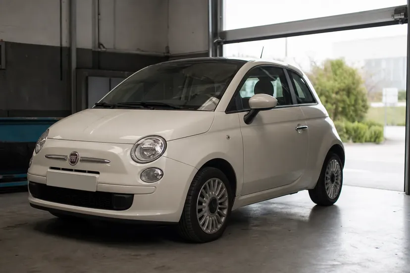 Une Fiat 500 thermique blanc crème vue de trois quarts avant, garée dans la travée d'un atelier devant une porte sectionnelle ouverte