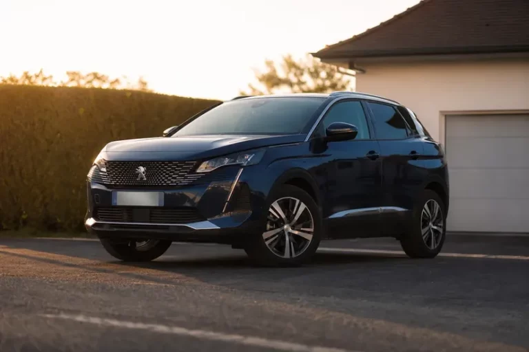 Un Peugeot 3008 de deuxième génération bleu foncé, vu de trois quarts avant en contre-plongée, arrêté sur l'allée bitumée d'une maison en fin de journée