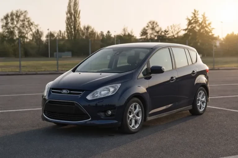 Ford C-Max bleu foncé vue de trois quarts avant, arrêtée moteur coupé sur le parking bitumé désert d'un terrain de sport, en fin de journée