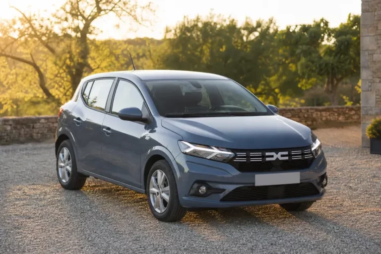 Une Dacia Sandero III bleu-gris vue de trois quarts avant, arrêtée dans une cour en gravier devant une maison de campagne en fin de journée.
