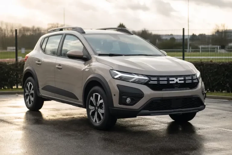 Une Dacia Sandero Stepway III beige sable vue de trois quarts avant, garée sur une aire de stationnement en enrobé humide au bord d'un terrain de sport, en milieu de matinée.