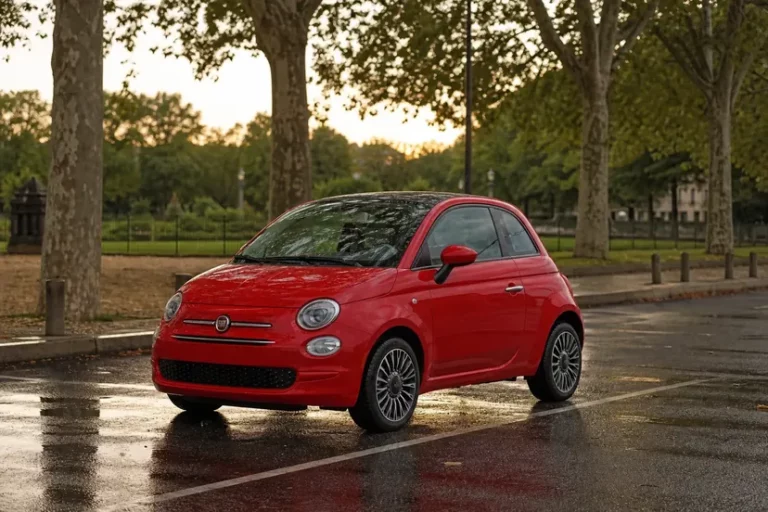Une Fiat 500 Hybrid rouge vue de trois quarts avant, garée en épi sur un parking bordé de platanes, sol mouillé, en fin de journée.