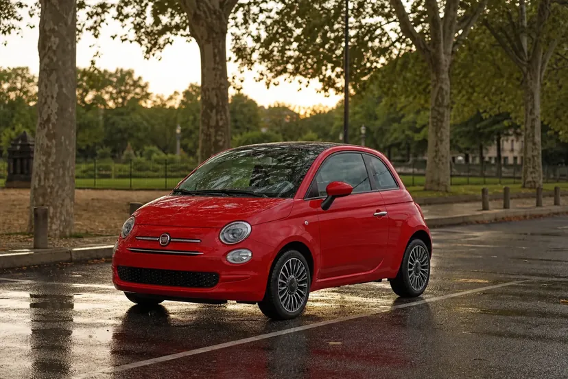 Une Fiat 500 Hybrid rouge vue de trois quarts avant, garée en épi sur un parking bordé de platanes, sol mouillé, en fin de journée.