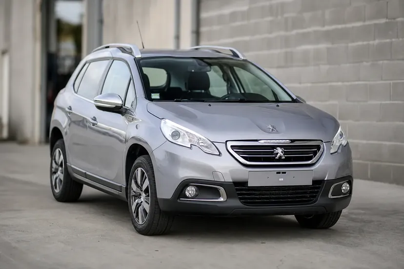 Une Peugeot 2008 de première génération gris argent, vue de trois quarts avant, à l'arrière d'un atelier.