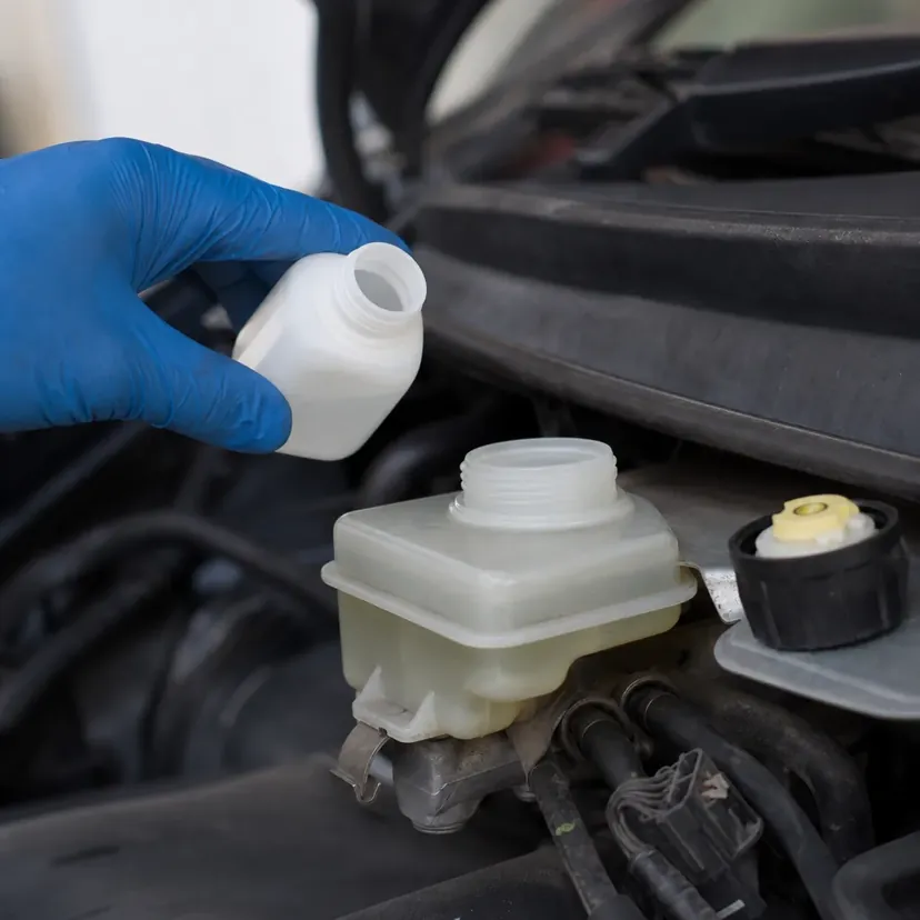 Gros plan sur le bocal de liquide de frein dans le compartiment moteur d'une citadine, bouchon dévissé, une main gantée approchant un bidon d'appoint.