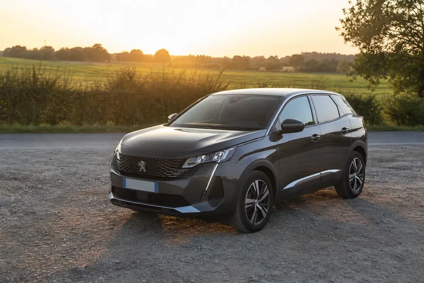 Un Peugeot 3008 de deuxième génération gris foncé, vu de trois quarts avant, arrêté sur une aire de stationnement en gravier au bord d'une route de campagne en fin de journée.