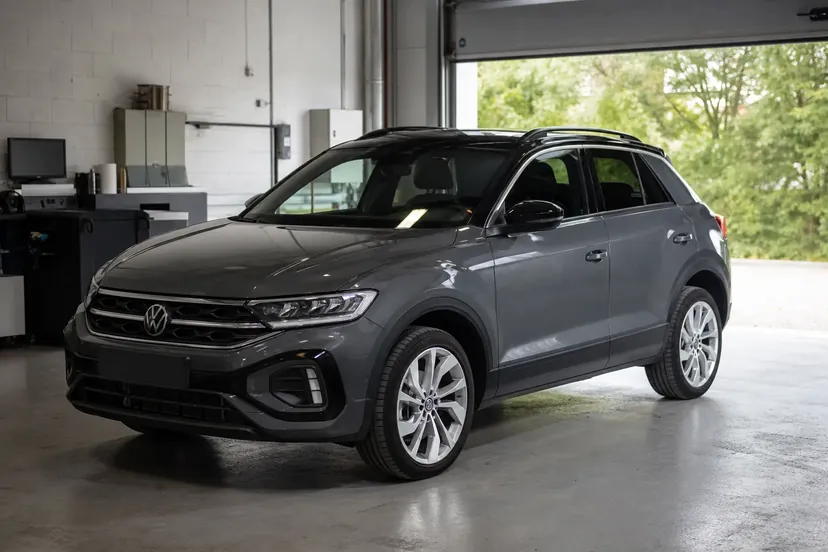 Volkswagen T-Roc gris foncé à toit noir, vu de trois quarts avant, garé dans la travée d'un atelier devant une porte sectionnelle ouverte.