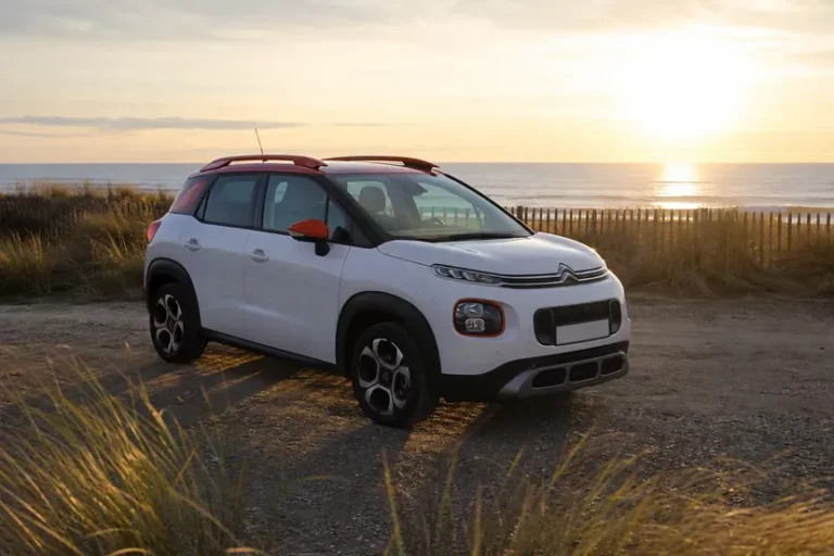 Un Citroën C3 Aircross blanc aux barres de toit et coques de rétroviseurs orange, vu de trois quarts avant, garé seul sur un parking de sable derrière une dune, face à l'océan en fin de journée.