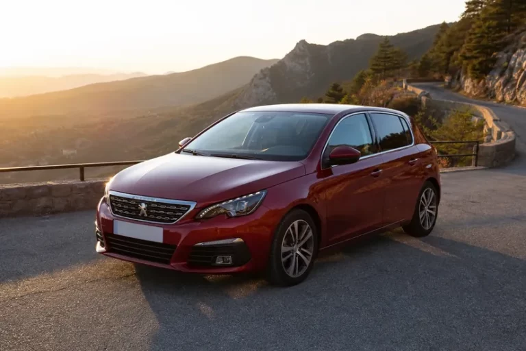 Une Peugeot 308 rouge foncé vue de trois quarts avant, garée seule sur un refuge au bord d'une route de montagne en lacets, en fin d'après-midi.