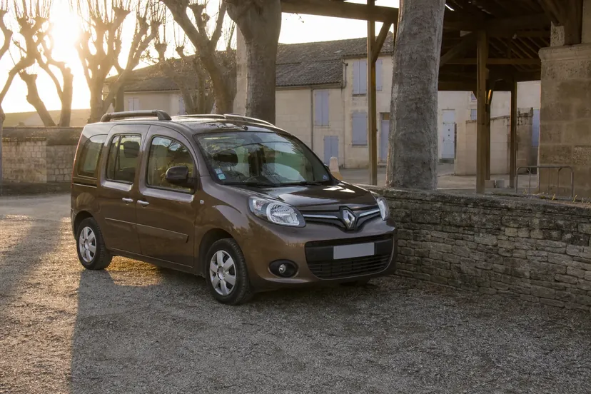 Un Renault Kangoo 2 brun doré métallisé vu de trois quarts avant, garé seul le long d'un muret devant une halle de village, en fin de journée d'hiver.